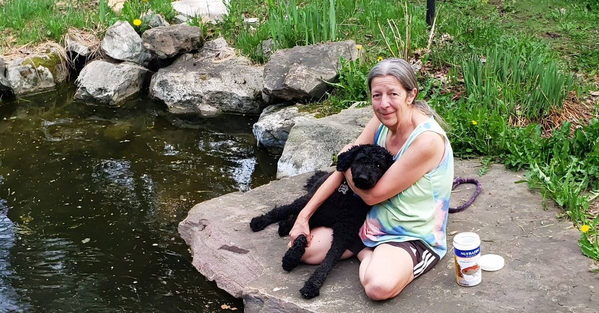 Jeannette sitting by a peaceful pond with her dog Sadie, surrounded by nature and soft greenery.
